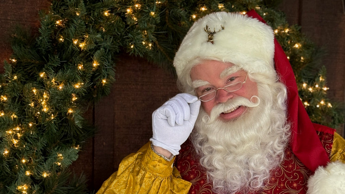 Santa Claus smiling in his traditional red hat and gold-trimmed suit beside an evergreen wreath decorated with white Christmas lights.