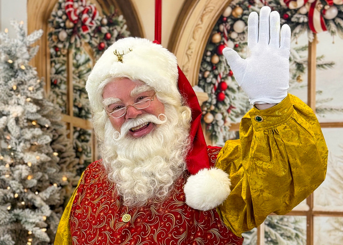 Santa Claus smiling and waving in a red and gold suit, surrounded by decorated Christmas trees, ornaments, and festive holiday greenery.